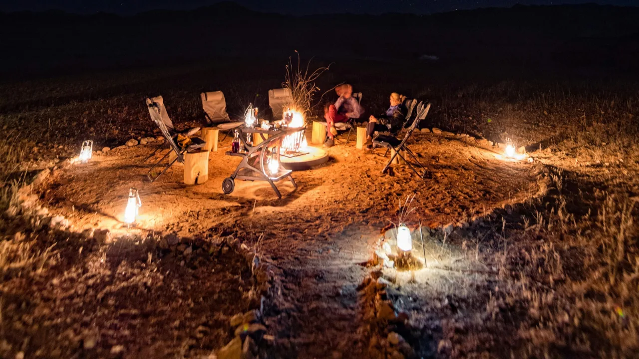 Evening desert safari camp in Dubai with tourists sitting around a bonfire, enjoying BBQ dinner and live entertainment under the night sky